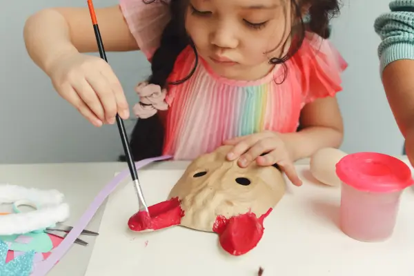Criança pintando uma máscara feita de papelão, em uma atividade de artesanato que estimula a criatividade, a expressão artística e o reaproveitamento de materiais simples.