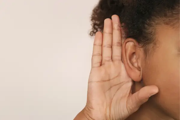 Criança coloca a mão no ouvido em gesto de escuta atenta, demonstrando participação em atividades sensoriais que estimulam a percepção auditiva e a atenção aos sons da natureza.