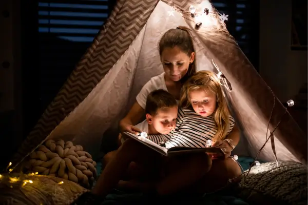 Mãe sentada dentro de uma barraca, contando histórias infantis para os filhos durante um acampamento ao ar livre, criando momentos mágicos em família sob a luz suave de uma lanterna.