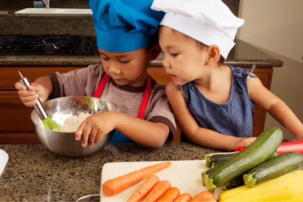 Duas crianças pequenas vestidas de chefs brincam de cozinhar em uma cozinha, misturando ingredientes em um bowl cercado por legumes coloridos — momento de gastronomia lúdica em família.