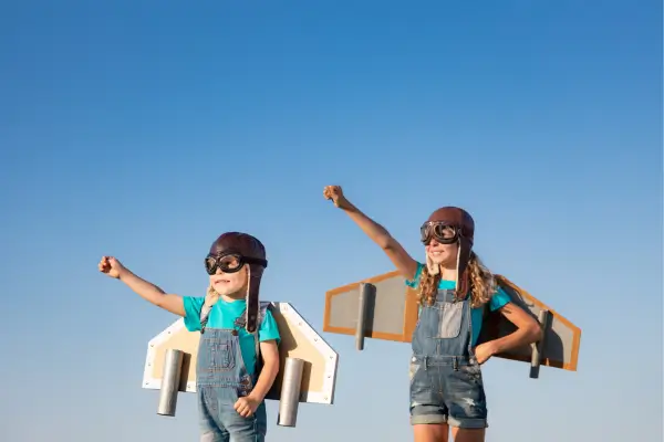 Crianças com aviões caseiros feitos de papelão nas costas brincam de voar, demonstrando os benefícios da criatividade e da imaginação nas atividades infantis ao ar livre.
