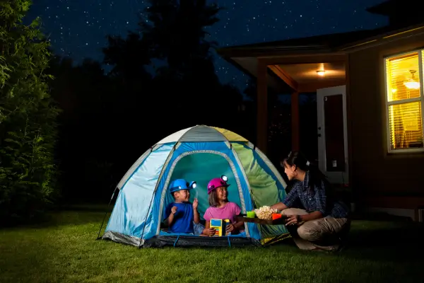 Crianças se preparam para observar o céu noturno durante um acampamento no quintal, com cobertores, lanternas e telescópio em um clima de curiosidade e descoberta ao ar livre.