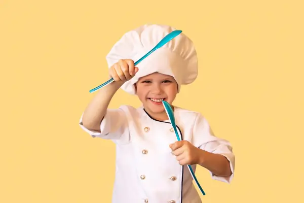 Criança vestida de chef com avental e chapéu branco, sorrindo enquanto participa de uma atividade de culinária ao ar livre.