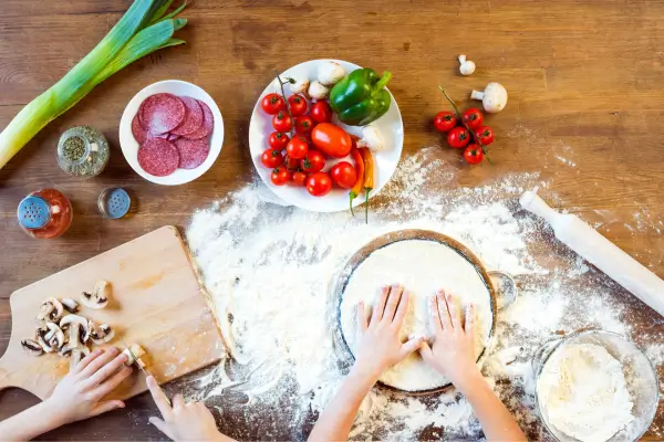 Criança preparando massa de pizza com as mãos, rodeada por ingredientes frescos, em uma experiência divertida de cozinha.