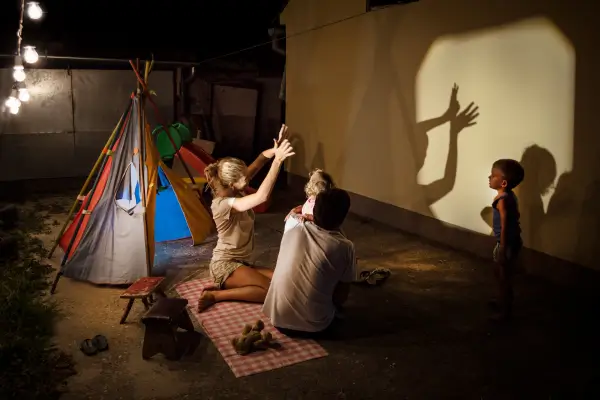 vCrianças se divertem em um acampamento no quintal brincando de teatro de sombras com uma lanterna e figuras recortadas, em um ambiente lúdico e criativo ao ar livre