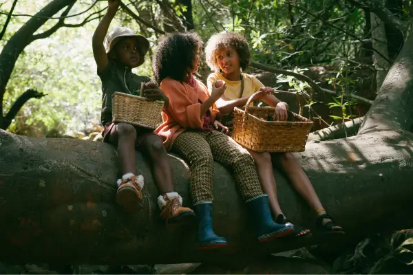 Crianças sentadas na floresta saboreiam frutas frescas de uma cesta, vivenciando atividades sensoriais que estimulam o paladar e promovem uma experiência de conexão com a natureza por meio dos alimentos.