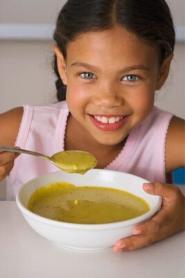 Menina em close tomando sopa ao ar livre no quintal, com expressão concentrada e aconchegante, segurando a tigela e com a colher na boca em um ambiente natural e acolhedor.
